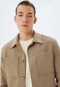 Tan jasje van zachte stof, met een knoopsluiting aan de voorkant, twee grote opgestikte zakken en een omgeslagen kraag, gedragen over een lichtgekleurde shirt.