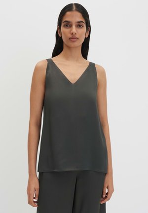 Femme aux longs cheveux foncés portant un haut sans manches gris foncé à col en V et un pantalon ample assorti, debout devant un fond clair uni.