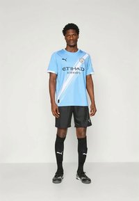 Lyseblå fotballdrakt med hvite detaljer og teksten "ETIHAD AIRWAYS", kombinert med svarte shorts, svarte sokker og svarte fotballsko.