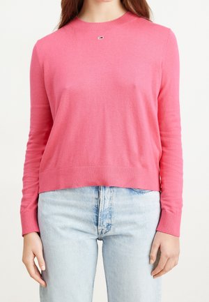 Žena v růžovém svetru s dlouhým rukávem a světle modrých džínách, stojící s uvolněnýma rukama podél těla na bílém pozadí.