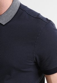 Polo shirt bleu marin avec un motif texturé à pois. Doté d'un col à carreaux blancs contrastant et de poignets élastiques.
