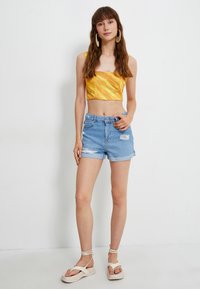 Top corto amarillo con textura acanalada, combinado con shorts de mezclilla azul claro con detalles desgastados. El modelo lleva sandalias en tonos neutros.