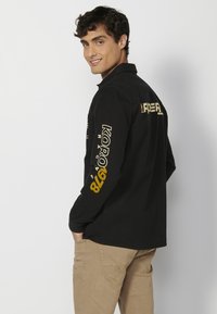 Camisa negra de manga larga que presenta gráficos de texto en amarillo y blanco en las mangas. El diseño incluye un cuello estándar y un ajuste slim.