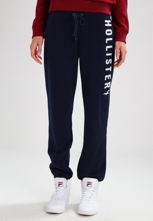 Personne portant un jogging bleu marine avec le texte "Hollister" sur le côté et des baskets montantes blanches Fila, debout devant un fond uni.