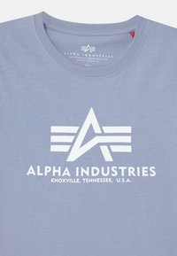 T-shirt de algodão azul claro com decote redondo. Apresenta uma impressão de logo branca com um emblema e o texto: "ALPHA INDUSTRIES, KNOXVILLE, TENNESSEE, EUA."