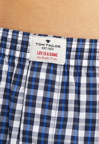 Tissu à carreaux bleu et blanc avec une taille élastique, présentant une étiquette avec "Tom Tailor" et le texte "La vie est un jeu, joue-le."