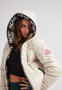 Giacca puffer bianca sporca con cappuccio foderato in logo nero, design trapuntato e un patch rosa sulla manica. Chiusura a zip e vestibilità cropped.