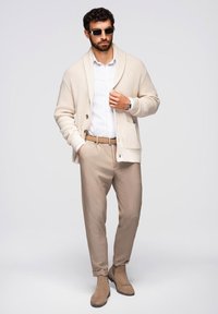 Beiger Strickcardigan über einem weißen Hemd, kombiniert mit hellbraunen Hosen und braunen Wildleder-Stiefeletten. Accessoires sind schwarze Sonnenbrillen.