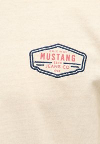 Tissu beige avec un logo cousu et imprimé indiquant "Original Mustang ESTD Jeans Co 1932" à l'intérieur d'un écusson bordé de bleu.