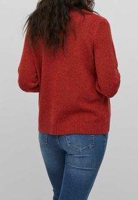 Czerwony sweter dziany z teksturowanym wykończeniem, ściągaczem u dołu i długimi rękawami, zestawiony z dżinsami. Widok od tyłu ukazuje luźny fason.