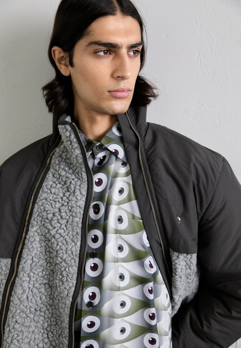 Herrejakke med et mørkegråt yderlag, fuzzy gråt indre og en mønstret skjorte med grønne, hvide og lilla øjemotiver.