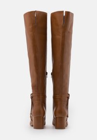 Bottes hautes jusqu'au genou en cuir marron avec un bout rond, un talon bloc, une fermeture éclair à l'arrière et des lanières décoratives à la cheville. Texture lisse et design épuré.