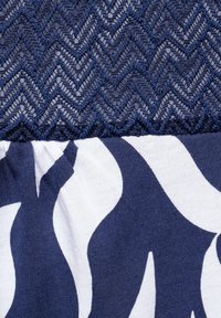 Marinblått tyg med en zigzag-mönstrad spets högst upp, som övergår till ett mjukt stretchmaterial med en vit abstrakt design nedanför.