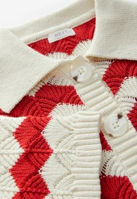 Polo en tricot avec un col crème et des motifs géométriques alternant entre rouge et crème. Dispose de deux grands boutons blancs au niveau du col.