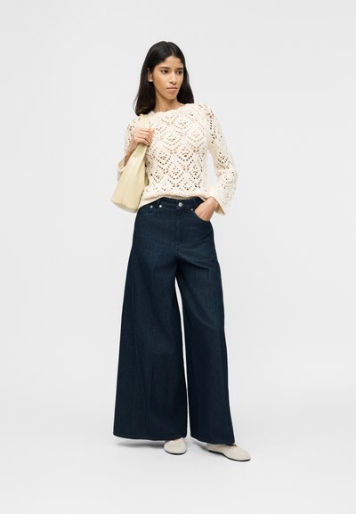 Γυναίκα που φοράει κρεμ πλεκτό πουλόβερ, σκούρα τζιν με φαρδιά πόδια, λευκά παπούτσια και κρατάει τσάντα ώμου σε κρεμ χρώμα, μπροστά σε απλό φωτεινό φόντο.