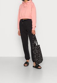 Růžová krátká mikina s kapucí a šňůrkou, černé tepláky, černé sandály na klínu a černá taška s abstraktními bílými liniemi.