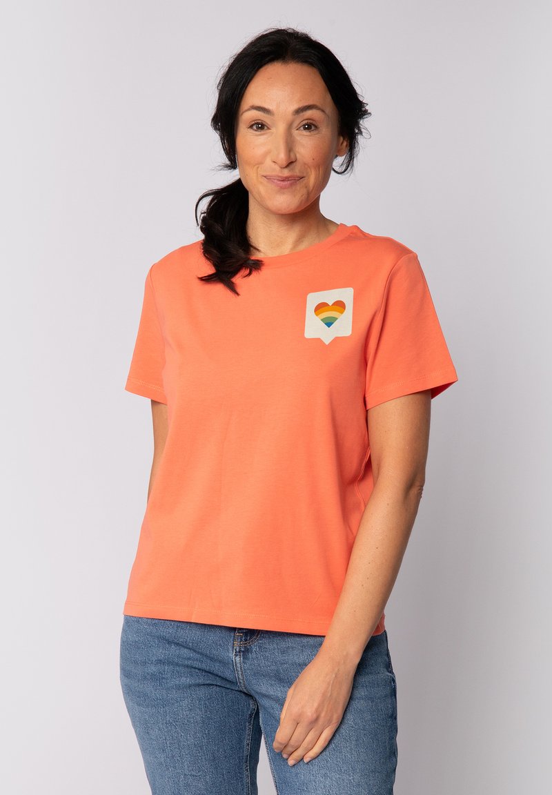 Koraal T-shirt van zachte stof, met een witte zakpatch met een regenboog hartontwerp. Korte mouwen en een relaxte pasvorm.