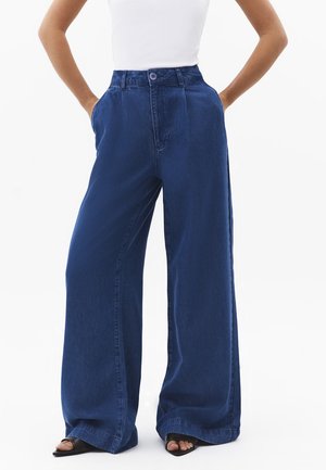 Frau trägt dunkelblaue, hochgeschnittene, weit geschnittene Jeans mit Knopfverschluss und Händen in den Taschen, kombiniert mit schwarzen offenen High-Heel-Schuhen.