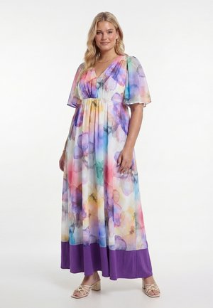 Frau trägt ein luftiges Maxikleid mit lila Saum, pastellfarbenem Blumenmuster, kurzen Puffärmeln und beigen Sandalen mit Absatz, steht vor weißem Hintergrund.