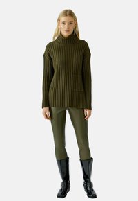Olivgrüner, gerippter Pullover mit hohem Kragen und Seitentasche, kombiniert mit enganliegenden olivgrünen Leggings und schwarzen kniehohen Stiefeln.