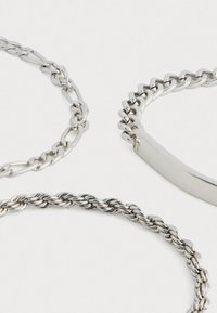 Három ezüst karkötő különböző mintákkal: az egyik egy lapos sáv, a másik egy csavart lánc, a harmadik pedig ovális láncszemekből áll.