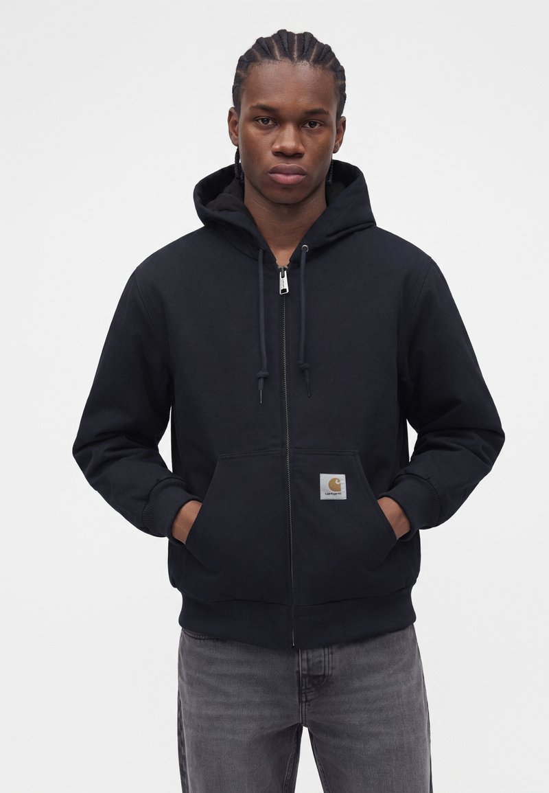 Joven con el pelo trenzado, vestido con una chaqueta negra con capucha y cremallera y vaqueros grises, de pie con las manos en los bolsillos de la chaqueta.