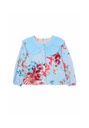 Junges Mädchen mit langen roten Haaren trägt ein blaues Kleid mit pinken und roten Blumenmustern und einem großen blauen Kragen, lächelnd mit der Hand auf der Hüfte.