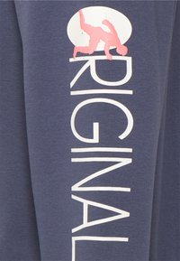 Sötétkék pulóver fehér "ORIGINAL" felirattal; a dizájn közepén egy kézállásban lévő személy rózsaszín grafikája található, kör alakú mintázatban.