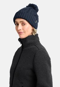 Berretto lavorato a maglia di colore blu navy con un pompon in cima, caratterizzato da un motivo testurizzato. Indossato con una giacca in pile nera, con la chiusura a zip visibile.