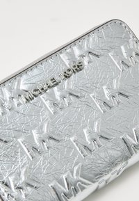 Billetera metálica plateada con patrón de logotipo en relieve "MK", hecha de material sintético texturizado, presenta un diseño elegante y cierre de cremallera.