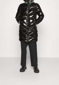 Svart, glänsande pufferjacka med hög krage och dragkedjestängning. Har ett chevronmönster och är matchad med lösa svarta byxor och robusta kängor.