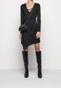 Schwarzes Wickelkleid mit langen Ärmeln, asymmetrischem Saum und V-Ausschnitt. Kombiniert mit knielangen schwarzen Lederstiefeln und einer Tasche mit Kettenakzent.