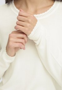 Pull blanc doux avec un col et des poignets en feston, doté d'une texture lisse et d'un tissu léger. Mains jointes devant.