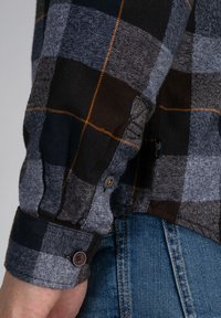 Camisa de cuadros negra y gris con acentos naranjas; presenta puños de botón y una textura suave. Primer plano de la manga y el área de la muñeca.