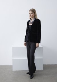 Černý sametový blazer s přiléhavým střihem, doplněný širokými černými kalhotami. Velký kulatý náhrdelník z korálků dodává detail. Minimalistické pozadí.