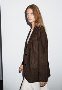 Brown suede blazer with two front pockets, two black buttons, and a notched collar. Model wears a light top and silver chain necklace.