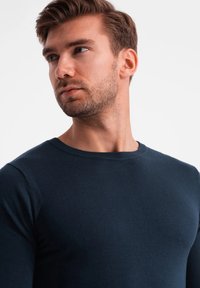 Mörkblå tröja med rund halsringning och en figurnära design. Slät struktur, långa ärmar och minimala sömmar. Modellen visar en sidoprofil.