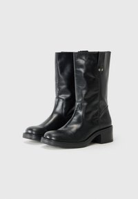 Botas de cuero negras hasta la rodilla con un tacón bajo, que presentan una superficie lisa y una correa lateral con un detalle de botón metálico.