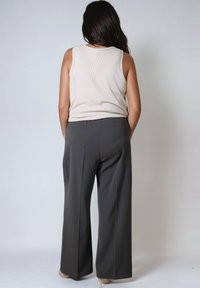 Femme aux longs cheveux noirs portant un top sans manches beige et un pantalon large gris foncé, se tenant les mains dans les poches, de dos.