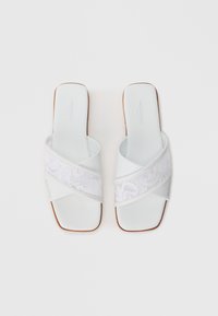 Hvite sandaler med glanset finish, med en crisscross snøringsrem og flate brune gummisåler. Merkenavnet er synlig på innersålen.