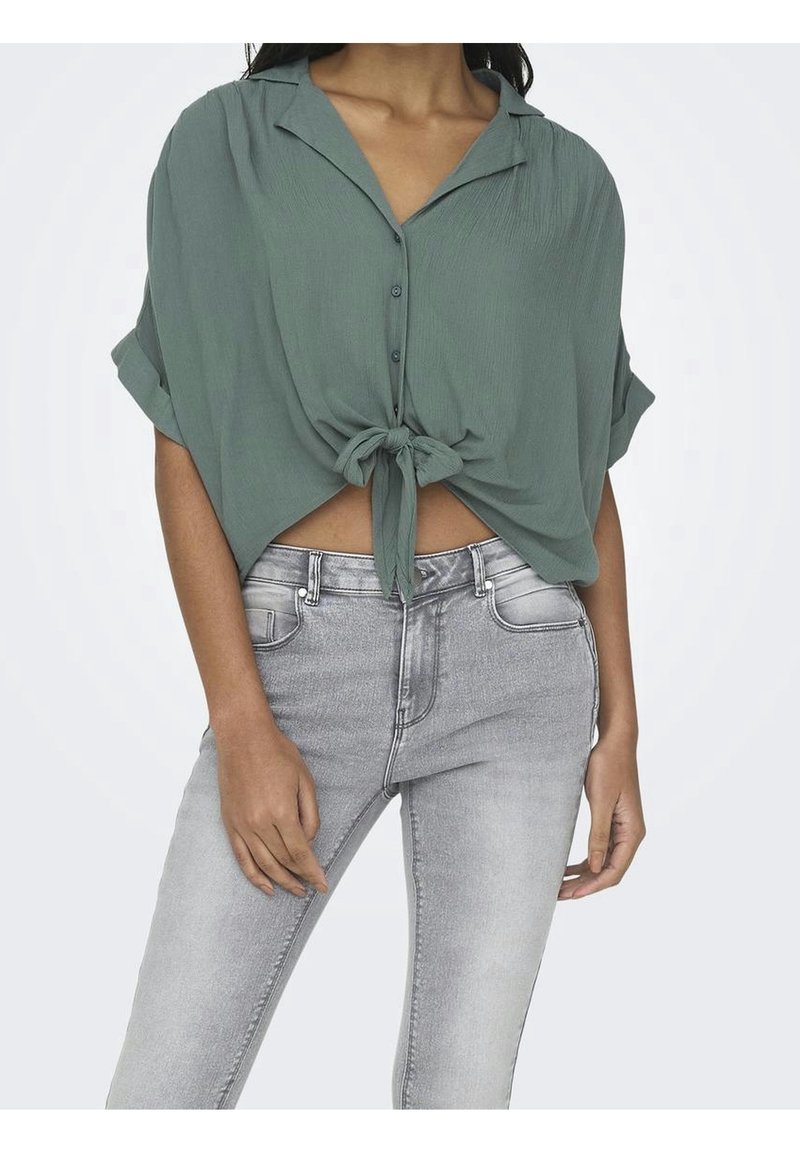 Blouse verte à manches courtes avec un devant boutonné, détail de noeud à la taille et tissu texturé, associée à un jean skinny gris clair.