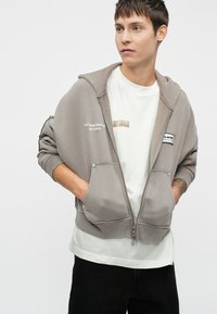 Halli hallatava lukuga hall kapuutsiga, millel on suured eesmiseks taskud ja tikitud tekst. Kantud valge T-särgi peal. Mustad püksid. Sile tekstuur ja mugav lõige.