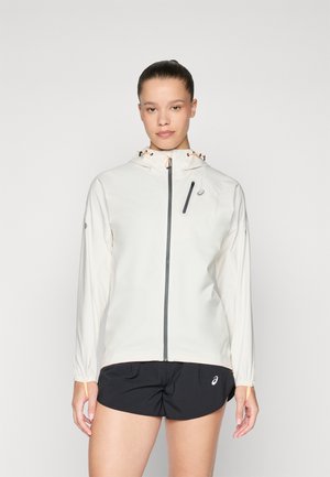 Vrouw in een witte sportjack met capuchon en rits aan de voorkant, met zwarte shorts met merkl logo's, staande tegen een effen achtergrond.