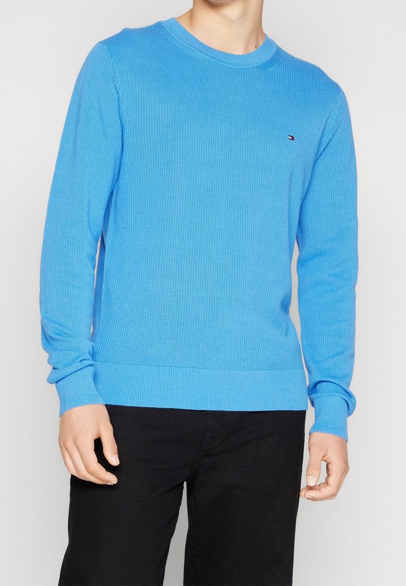 Uomo che indossa un maglione a maniche lunghe di lana blu brillante con un piccolo logo sul petto e pantaloni neri, su uno sfondo semplice.