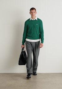 Pulover verde cu textură canalată peste o cămașă albă, pantaloni gri și pantofi negri. Poartă o geantă tote neagră, mergând pe un pardoseală neutr�ă.