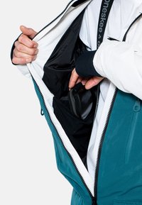 Türkis-weiße Jacke mit Reißverschluss vorne; Innentasche sichtbar, hält einen schwarzen Gegenstand; glattes, flexibles Material; verstellbare Ärmelbündchen.