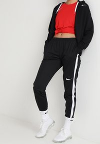 Pantalones de chándal negros con rayas laterales blancas y un logo icónico, combinados con una camiseta corta roja y una sudadera negra, zapatillas blancas.