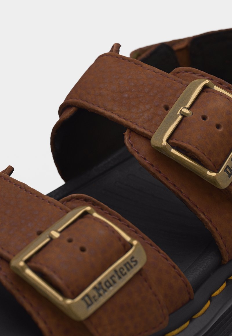 Braune Ledersandalenriemen mit goldfarbenen Metallschnallen, die mit "Dr. Martens" beschriftet sind, auf einer schwarzen Sohle mit gelber Naht.