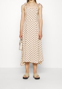 Vestido midi sin mangas en color beige con manchas marrones, tirantes ajustables y un dobladillo asimétrico. Combinado con sandalias deslizantes beige y un bolso pequeño.