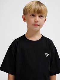 Blond chłopiec z fryzurą na kształt miski, ubrany w czarną koszulkę z małym białym logo pszczoły na piersi, patrzący lekko na bok.